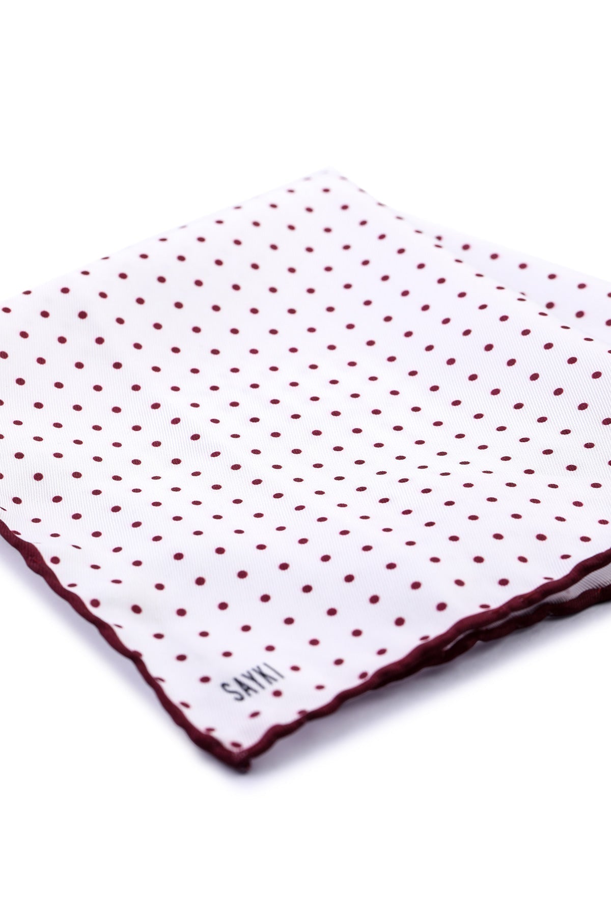 White pocket square with burgundy polka dots and a matching scalloped edge, featuring the brand name ’saiki’ embroidered in black at one corner.