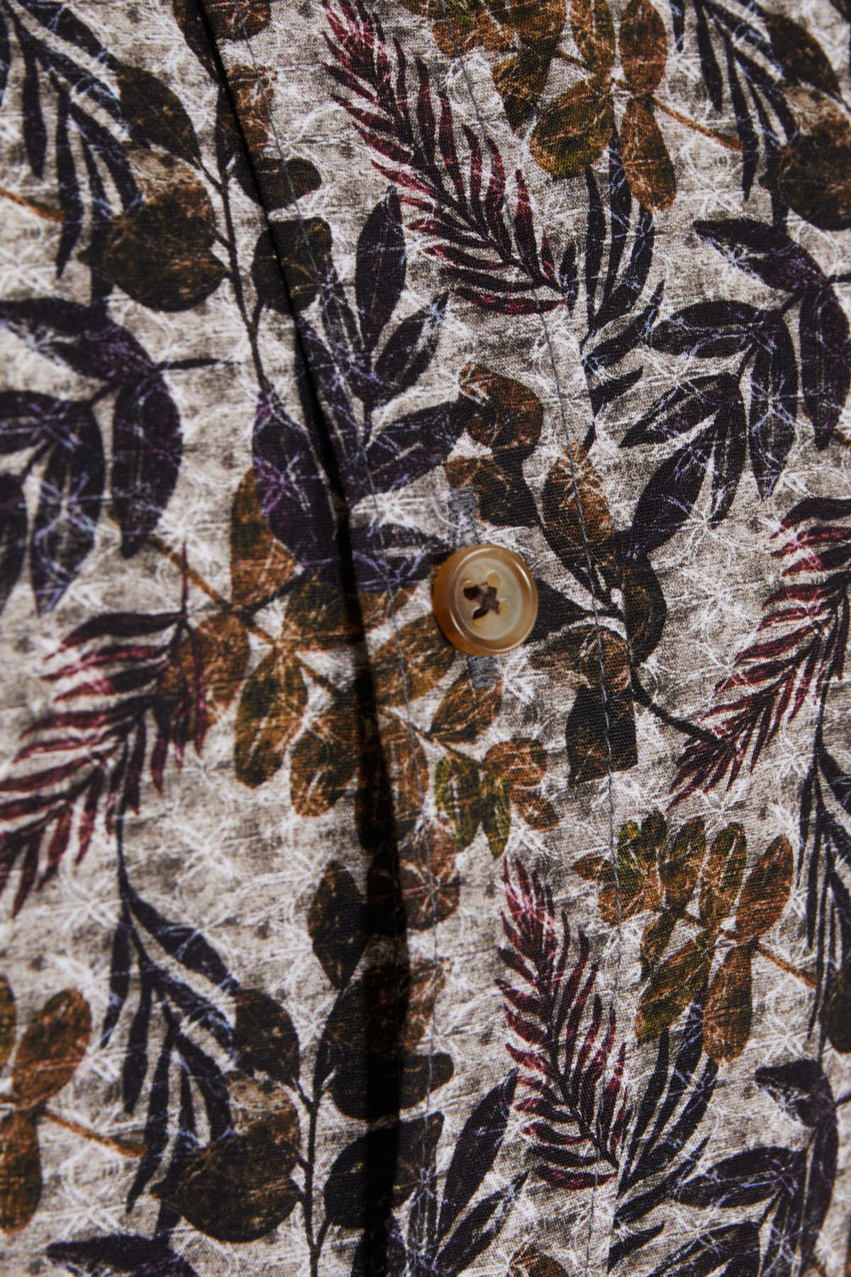 A beige button with a subtle cross design sits against a fabric featuring an intricate leaf pattern in shades of brown, black, and muted purple.