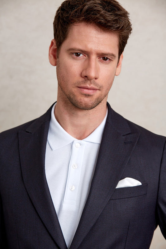 A man wearing a dark navy suit jacket over a light blue textured polo shirt with white buttons and a crisp white pocket square.