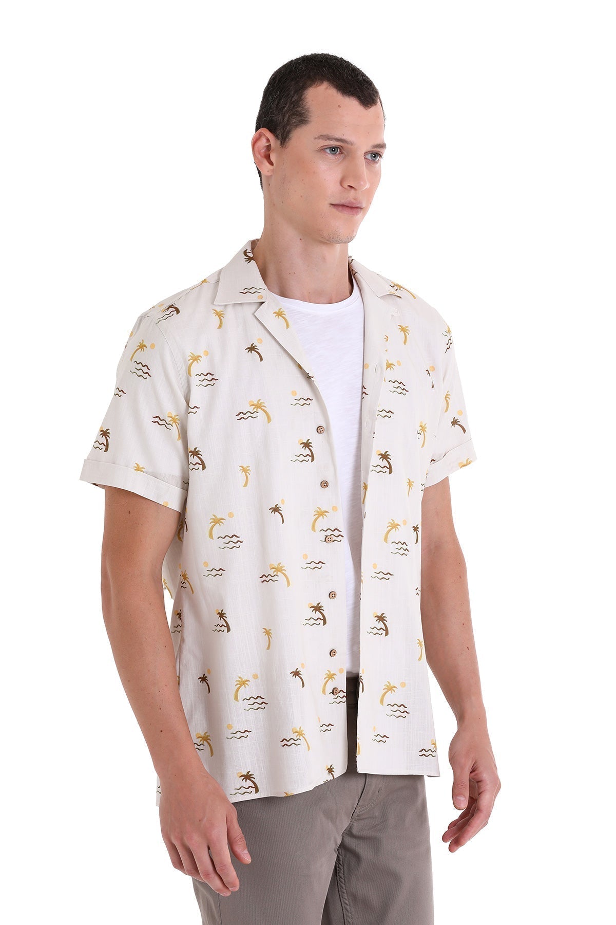 A man wearing a short-sleeved, button-up shirt with a tropical print of palm trees and wave patterns in gold and dark brown on an off-white fabric.
