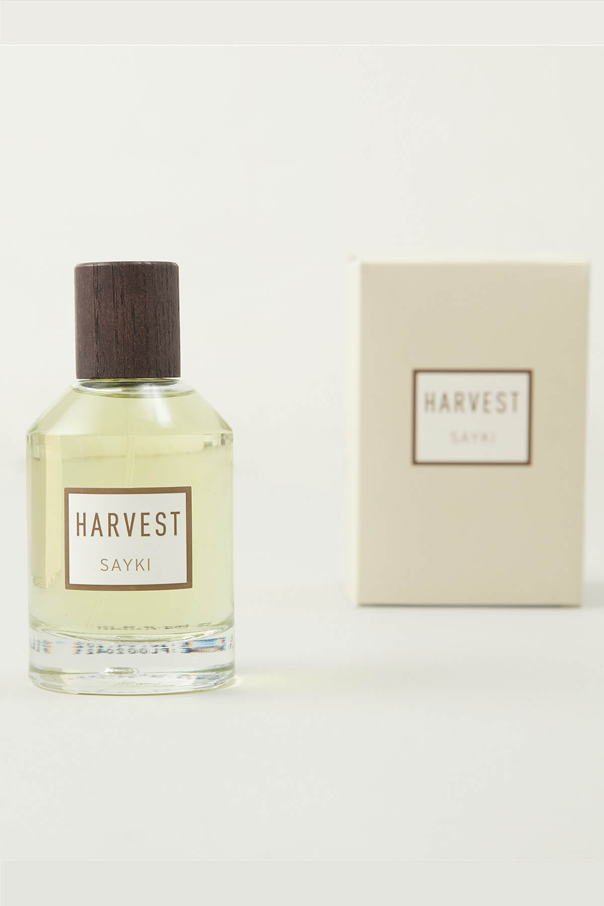 A clear glass bottle of sayki harvest men’s eau de parfum 100 ml with dark wood cap and pale yellow liquid beside cream box
