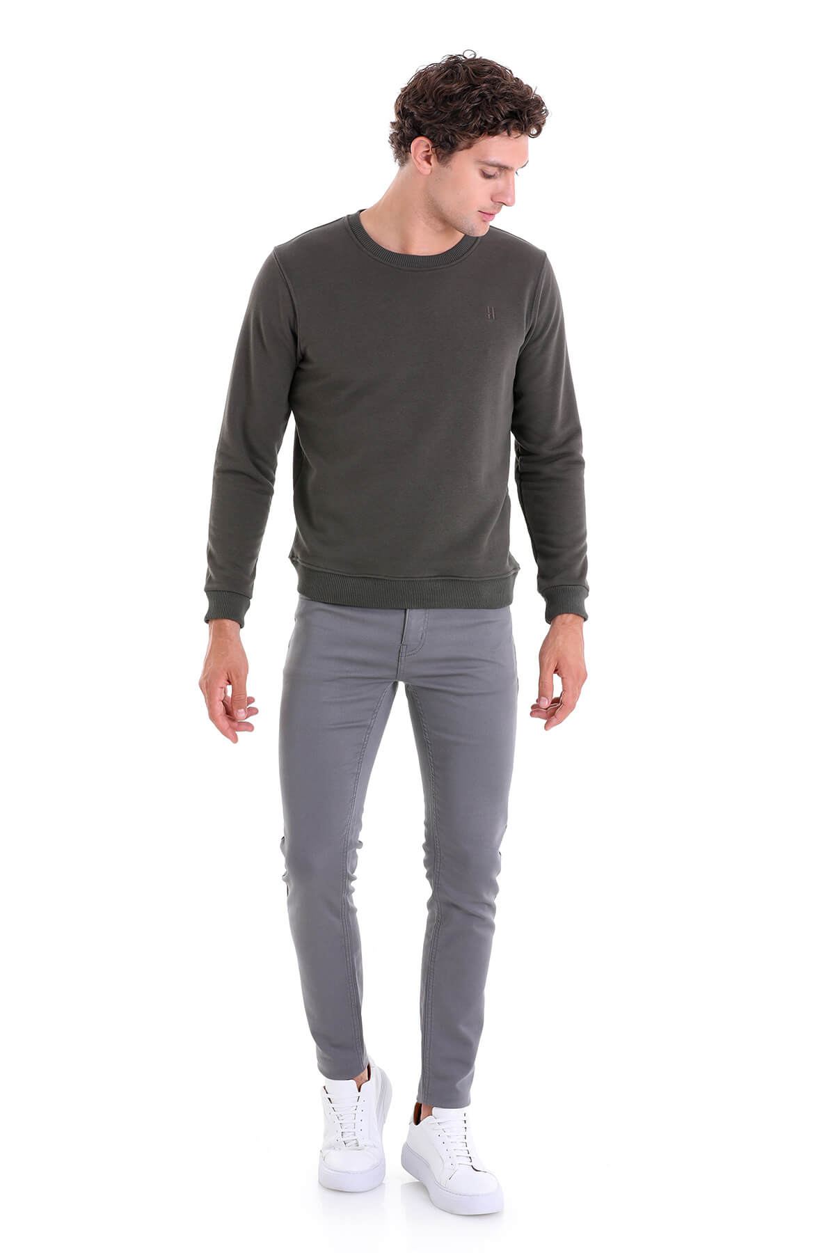 A man wearing a dark olive green long-sleeved crewneck sweatshirt paired with slim-fit gray trousers and white sneakers.