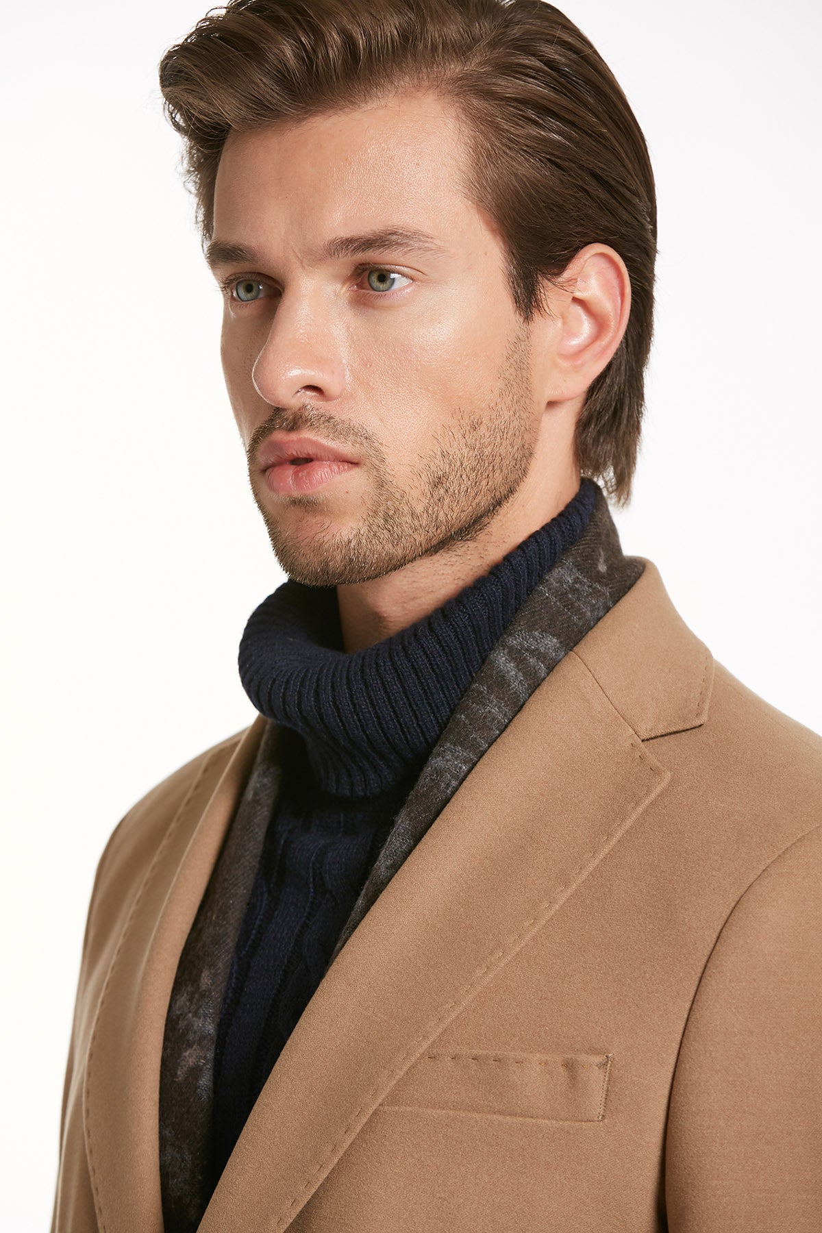 A tan wool overcoat with a notched lapel worn over a dark blue ribbed turtleneck and a patterned scarf.