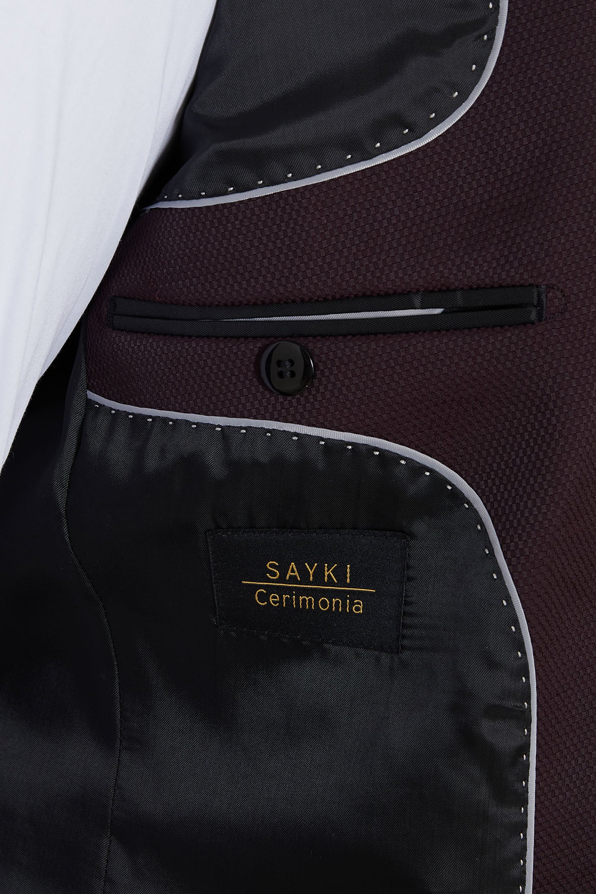 A black satin interior lining with silver stitching and a gold-embossed ’sayki cerimonia’ label on the inside of a maroon textured suit jacket.