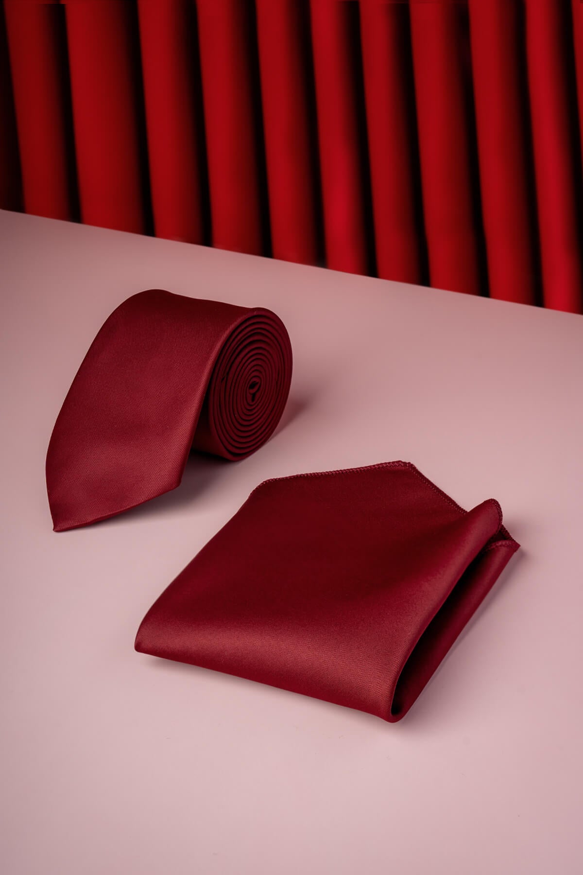 A rich burgundy silk tie and matching pocket square set, elegantly arranged on a soft pink surface.