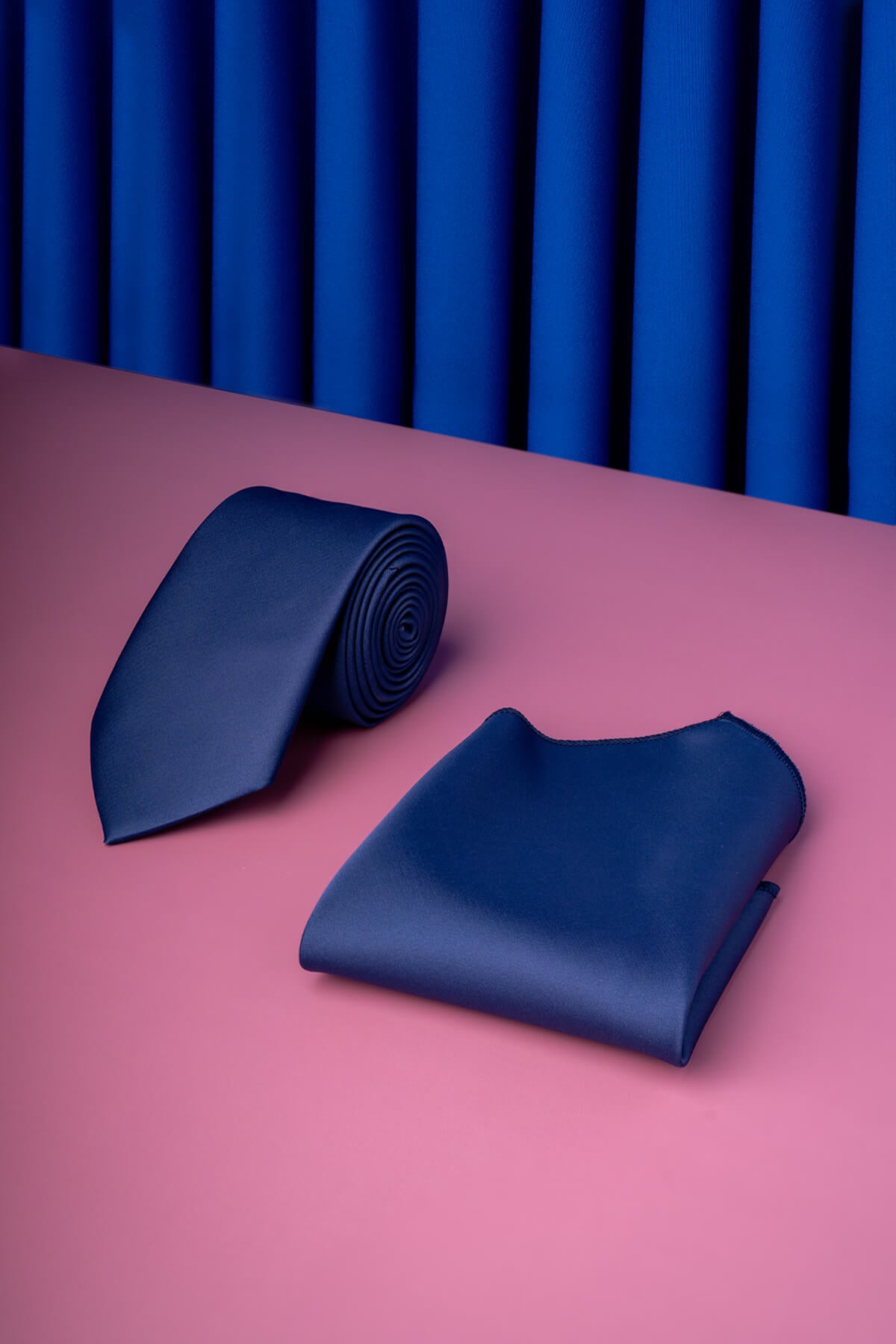 A sleek navy blue satin tie and matching pocket square, elegantly arranged on a pink surface.