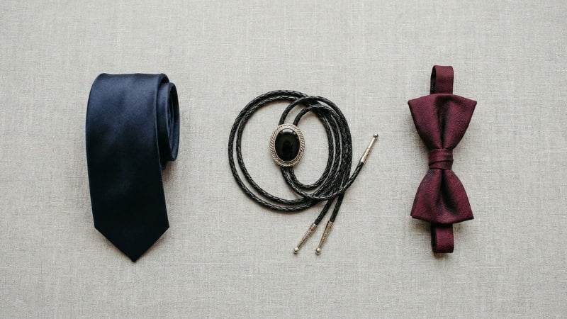 Flat lay comparison of a navy necktie, silver and onyx bolo tie, and burgundy bow tie