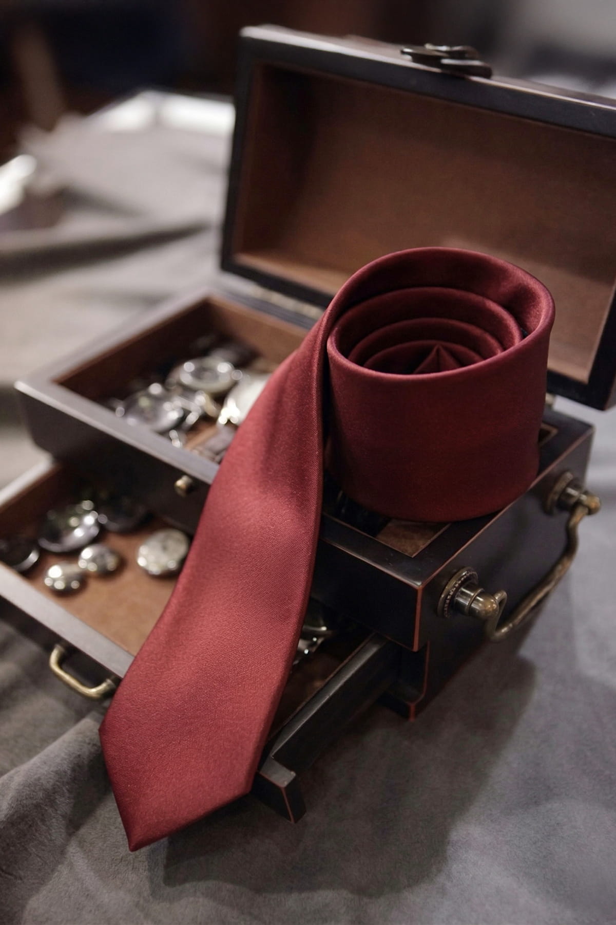 A rich burgundy silk casual 2.36 inches burgundy tie coiled on a dark wooden chest with brass hardware