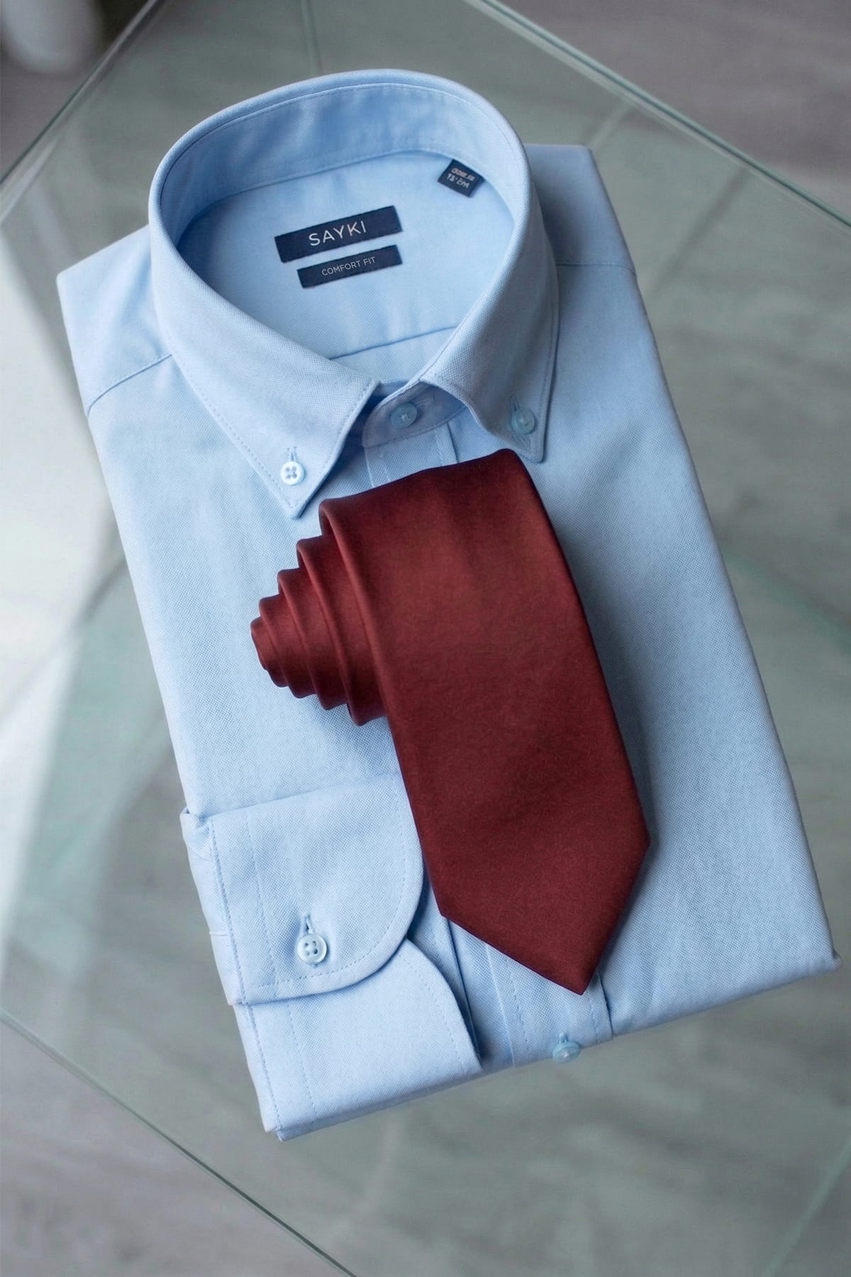 A neatly folded light blue button-down shirt with sayki label paired with a casual 2.36 inches burgundy micro polyester tie