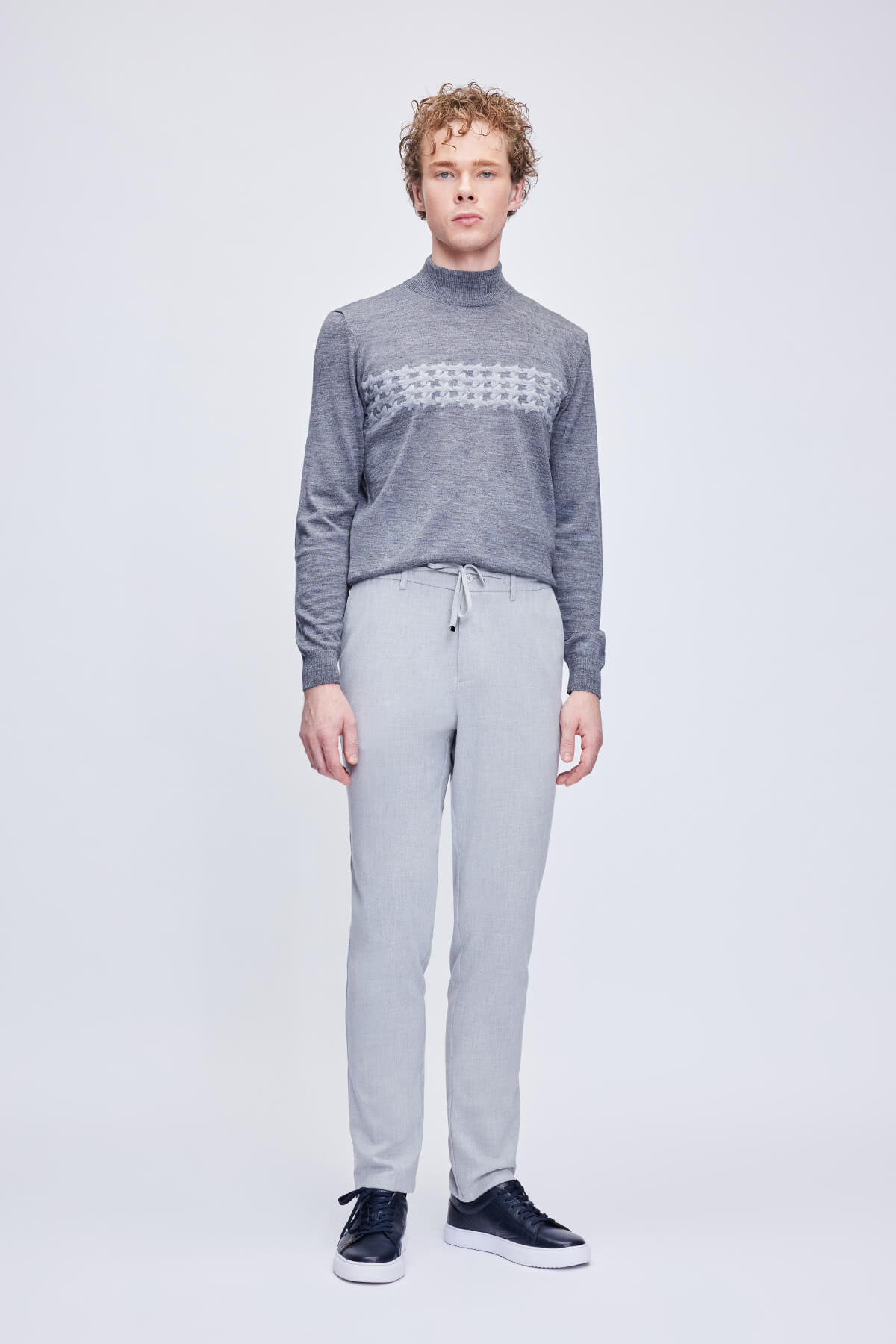 A man wearing a gray textured turtleneck sweater with a white geometric pattern across the chest and light gray tapered trousers.