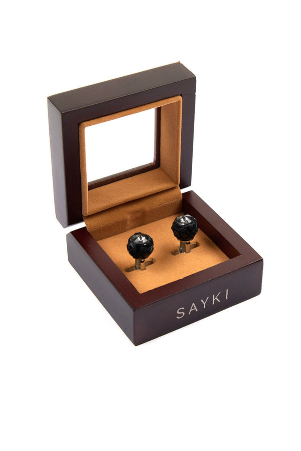 Black, faceted spherical cufflinks with metallic stems nestled in a dark wood box lined with tan velvet.
