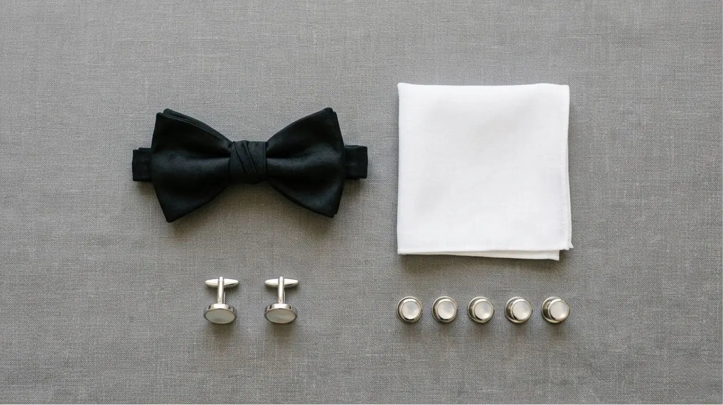 Overhead flat-lay of a black self-tie bow tie, silver studs, cufflinks, and a folded white pocket square arranged on a muted gray surface, no model
