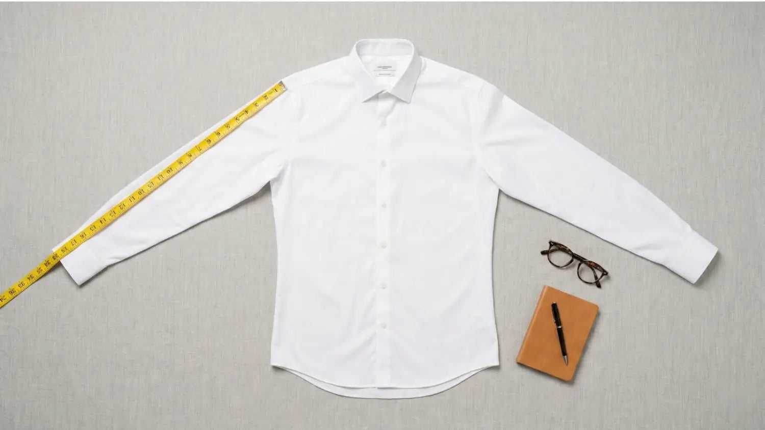 White dress shirt laid flat with a yellow measuring tape along the sleeve, a notebook, pen, and glasses on a gray surface