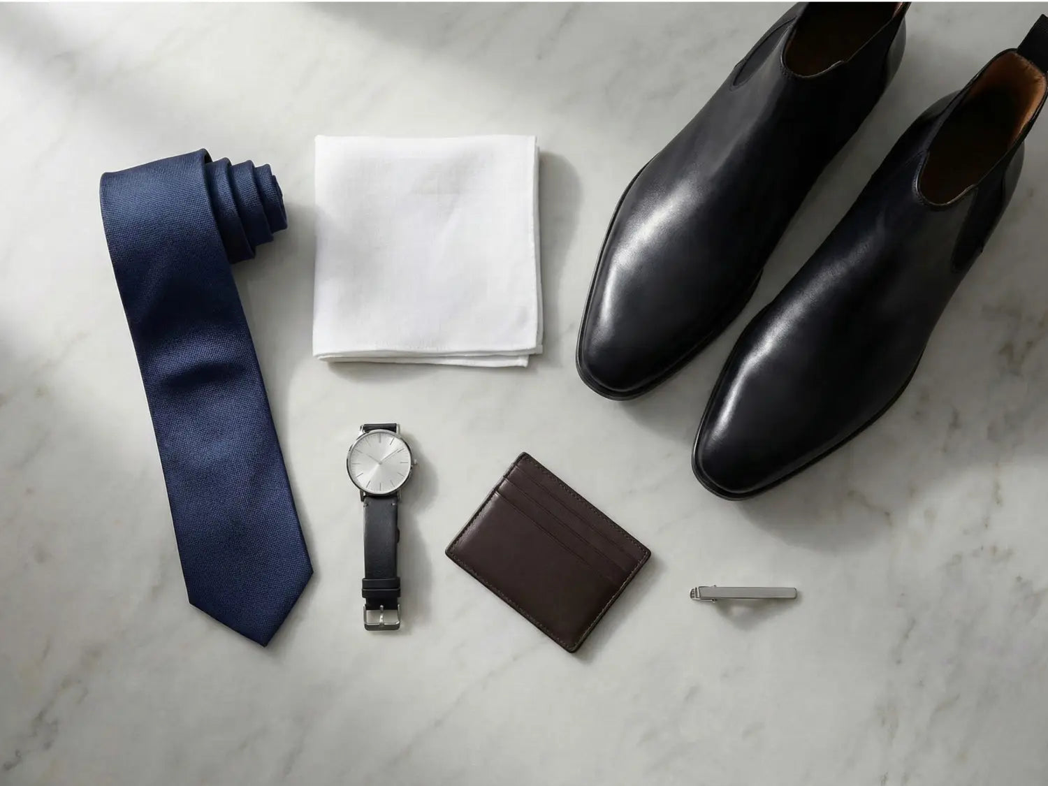 Flat lay of men’s suit accessories including navy tie, white pocket square, black leather dress shoes, minimalist watch, brown leather wallet, and silver tie bar arranged on a marble surface.