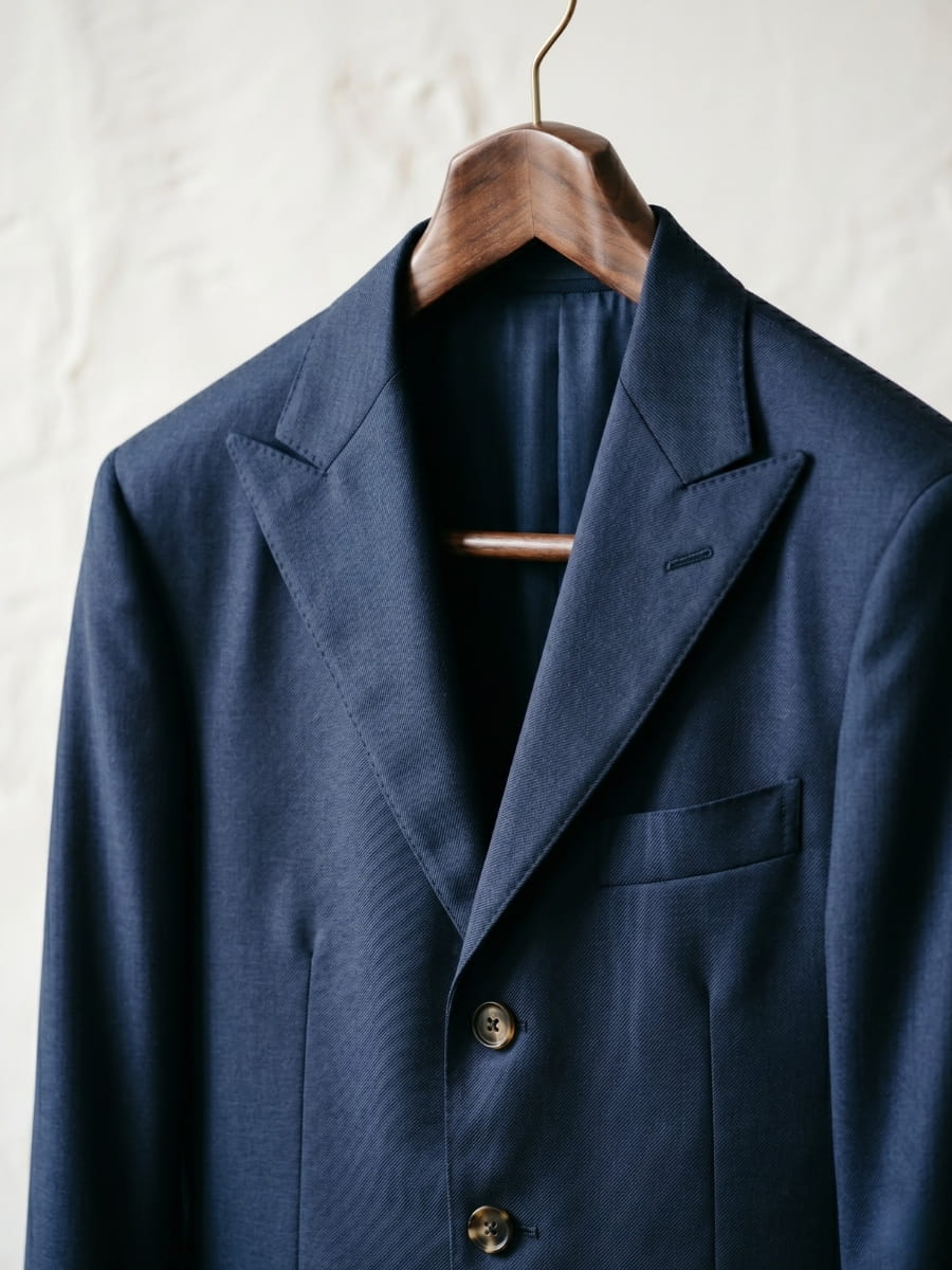 Close-up of navy worsted wool suit jacket on wooden hanger showing smooth refined diagonal twill weave with peak lapel and chest pocket detail