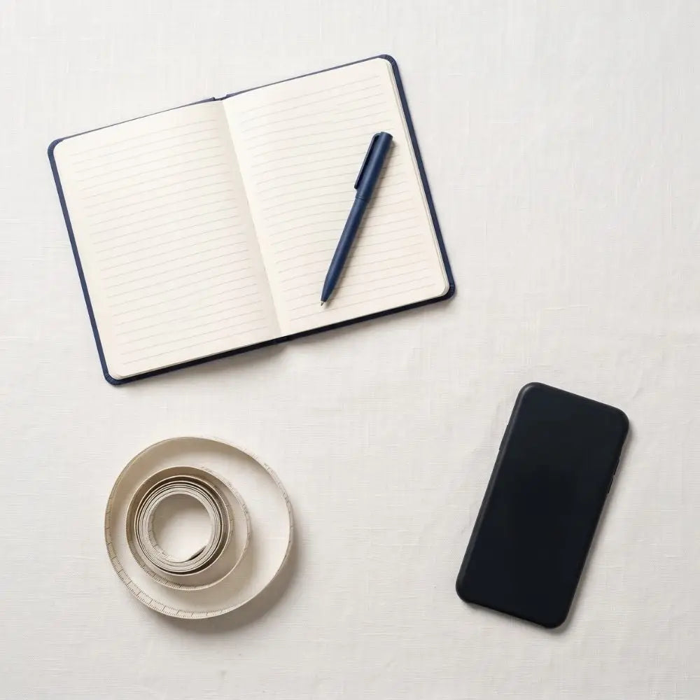 Open notebook, pen, coiled tape measure, and phone laid out for recording suit measurements