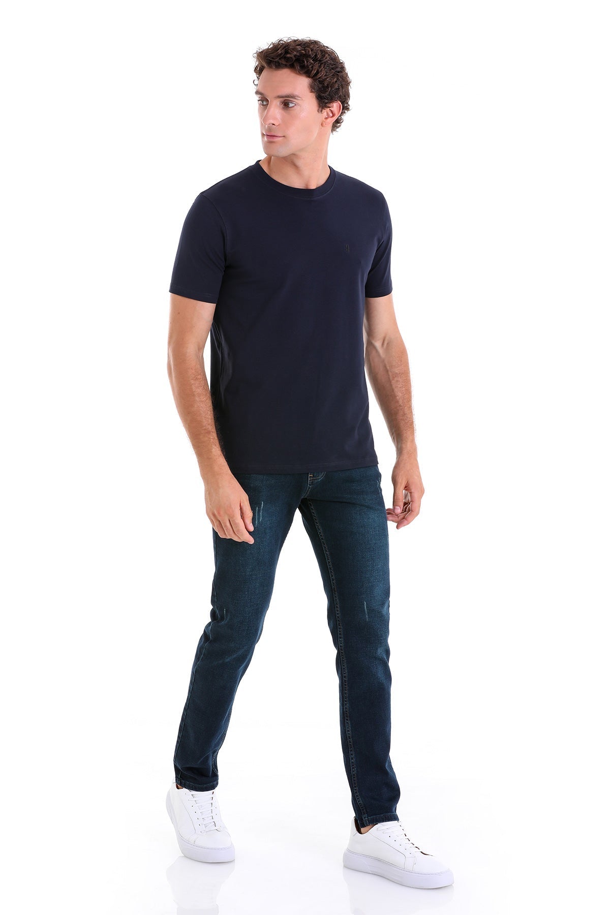 A man wearing a dark navy short-sleeved crew neck t-shirt paired with slim-fit dark blue jeans and white sneakers.