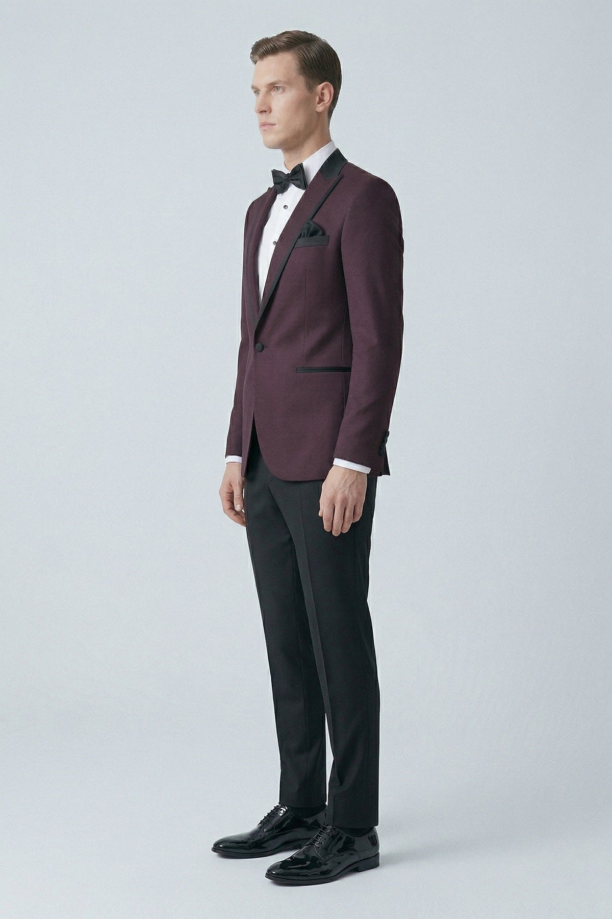 A man wearing a slim fit classic tuxedo with a burgundy peak lapel jacket and black satin trim and trousers