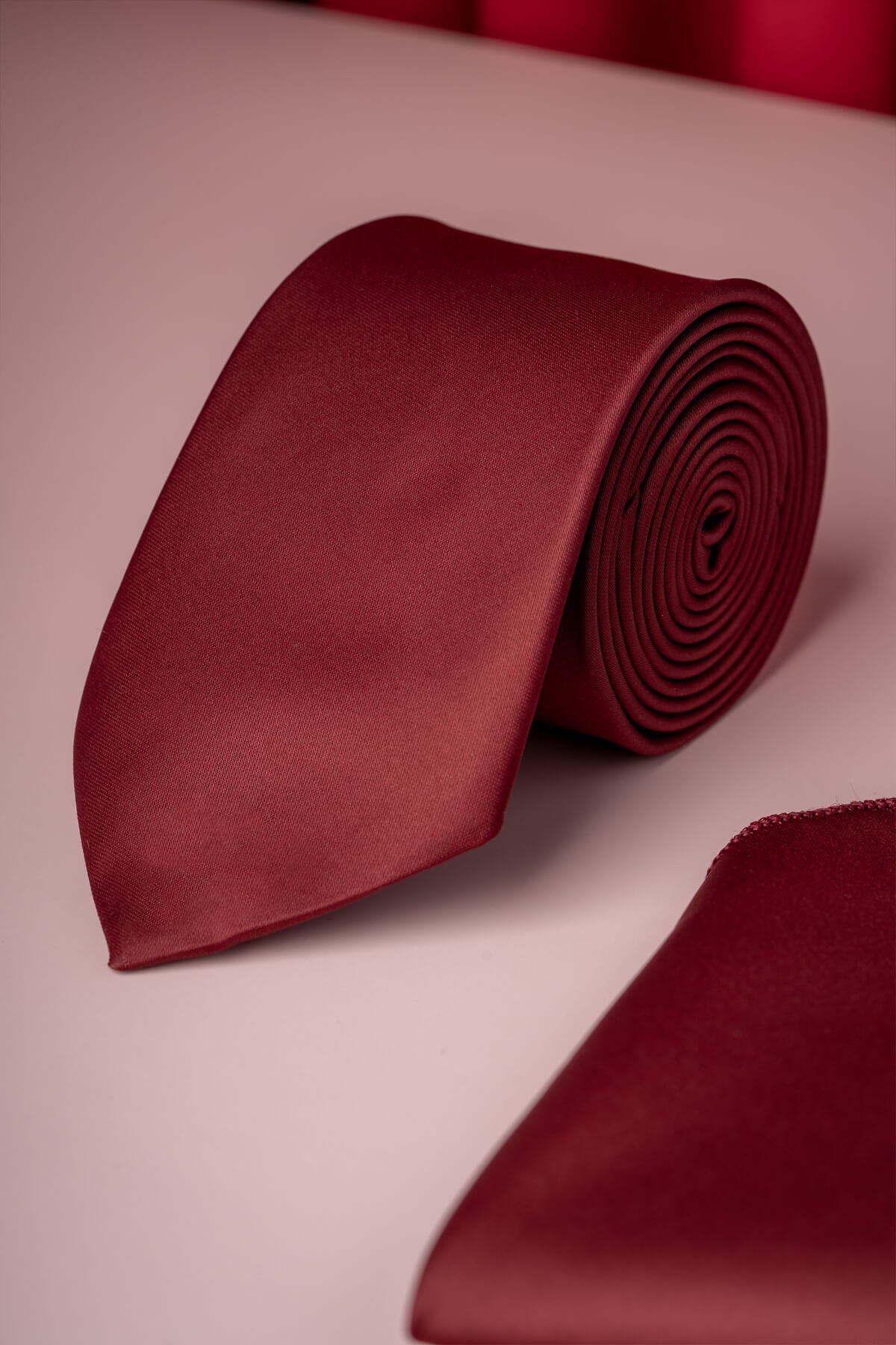 A rich burgundy satin tie, neatly rolled and displayed alongside a matching pocket square.