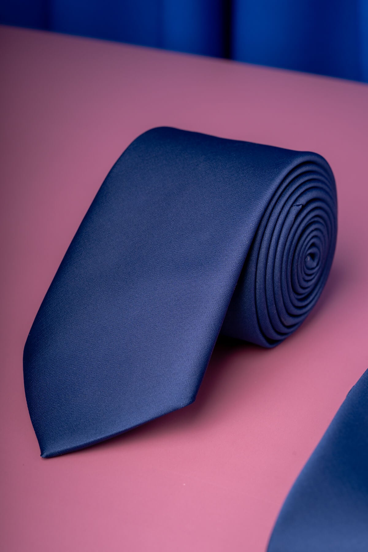 A sleek navy blue satin tie, neatly rolled and displayed on a soft pink surface.