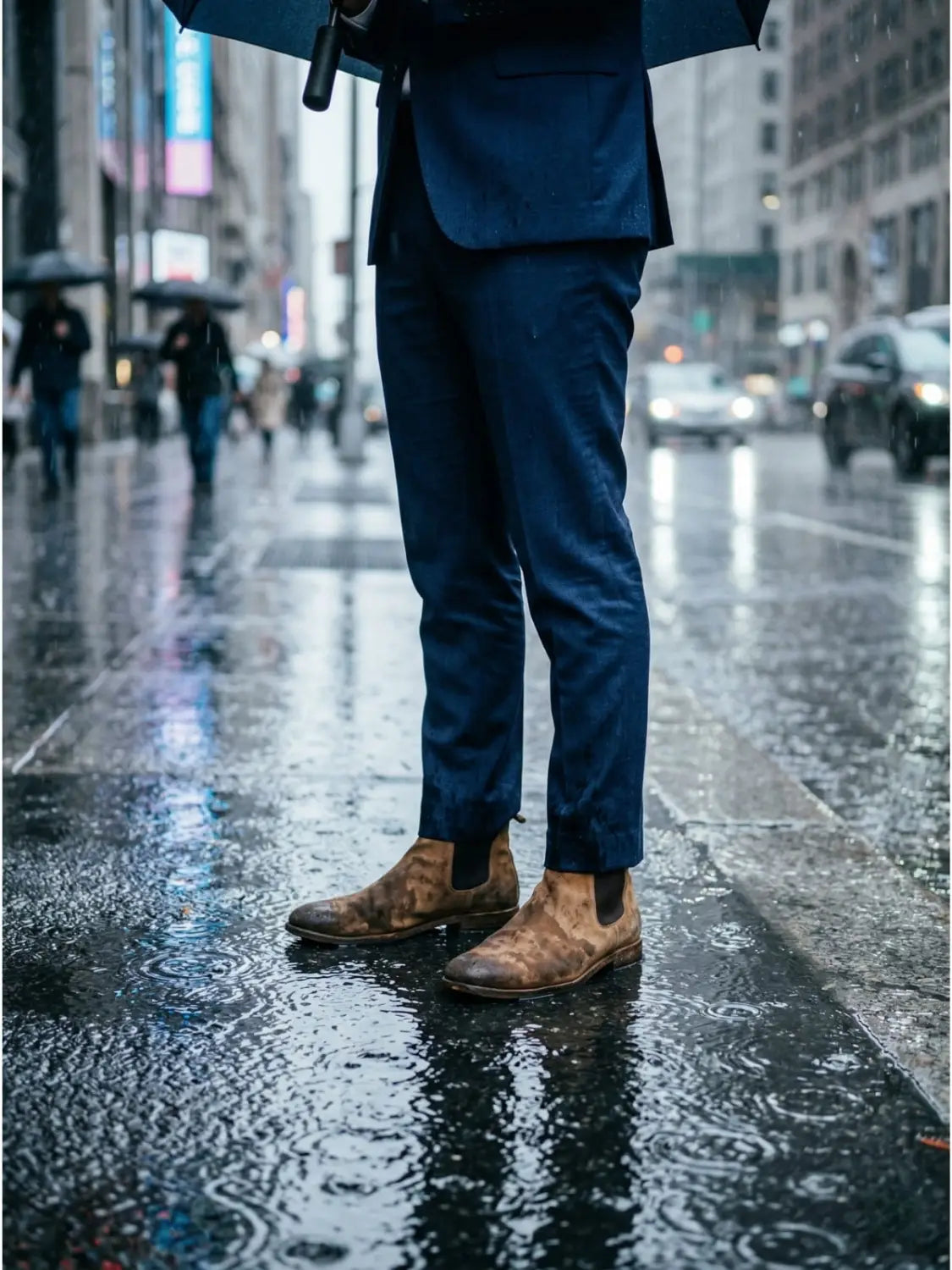 Man in a navy suit standing on a rain-soaked city sidewalk with water-stained tan suede Chelsea boots showing visible moisture damage