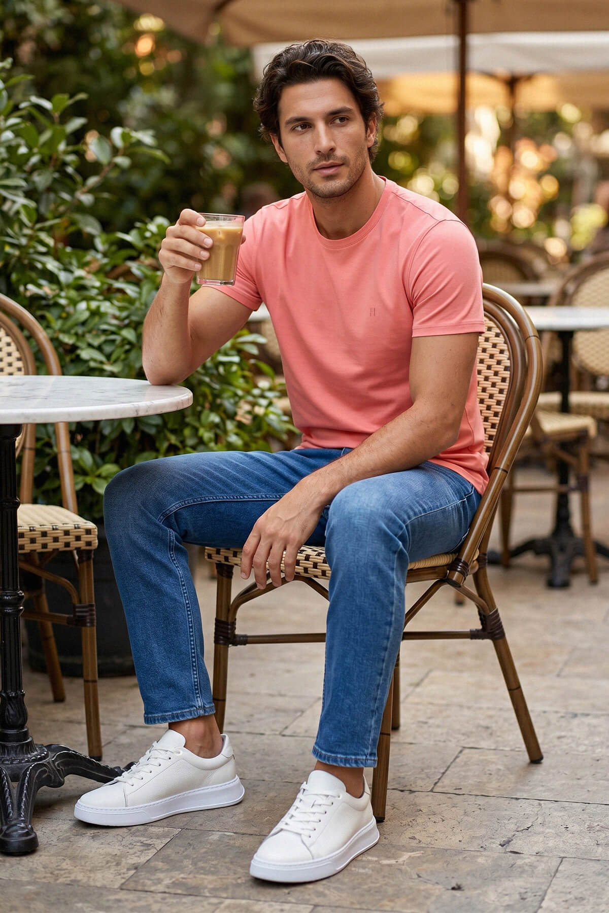 A man wearing white pebbled leather lace up sneakers seated outdoors in coral pink t shirt and blue jeans